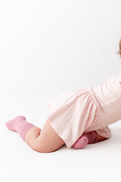 A baby wearing a light pink short-sleeve romper and matching pink ribbed socks, kneeling on a white studio floor.