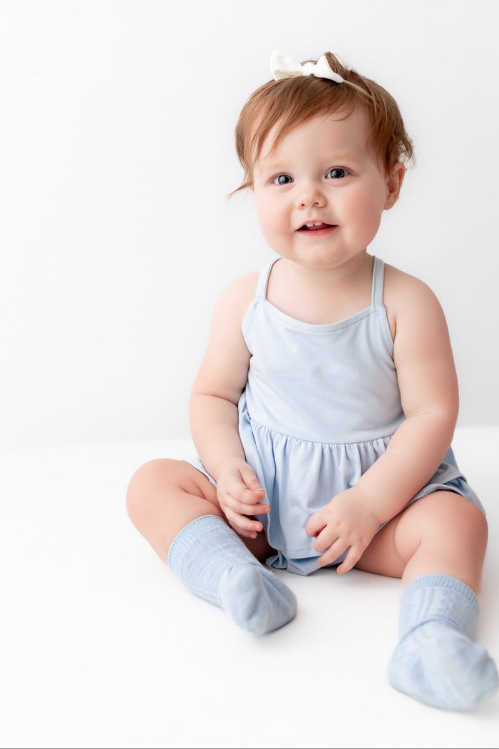 A baby girl sitting on a white studio floor wearing a light blue sleeveless romper and matching socks, with a small white bow in her hair. An adult hand is pointing from the right side.