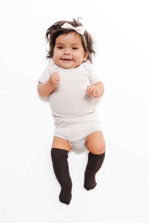 A smiling baby lying on a white background, wearing a light gray short-sleeve bodysuit, dark brown knee-high socks, and a white bow headband.