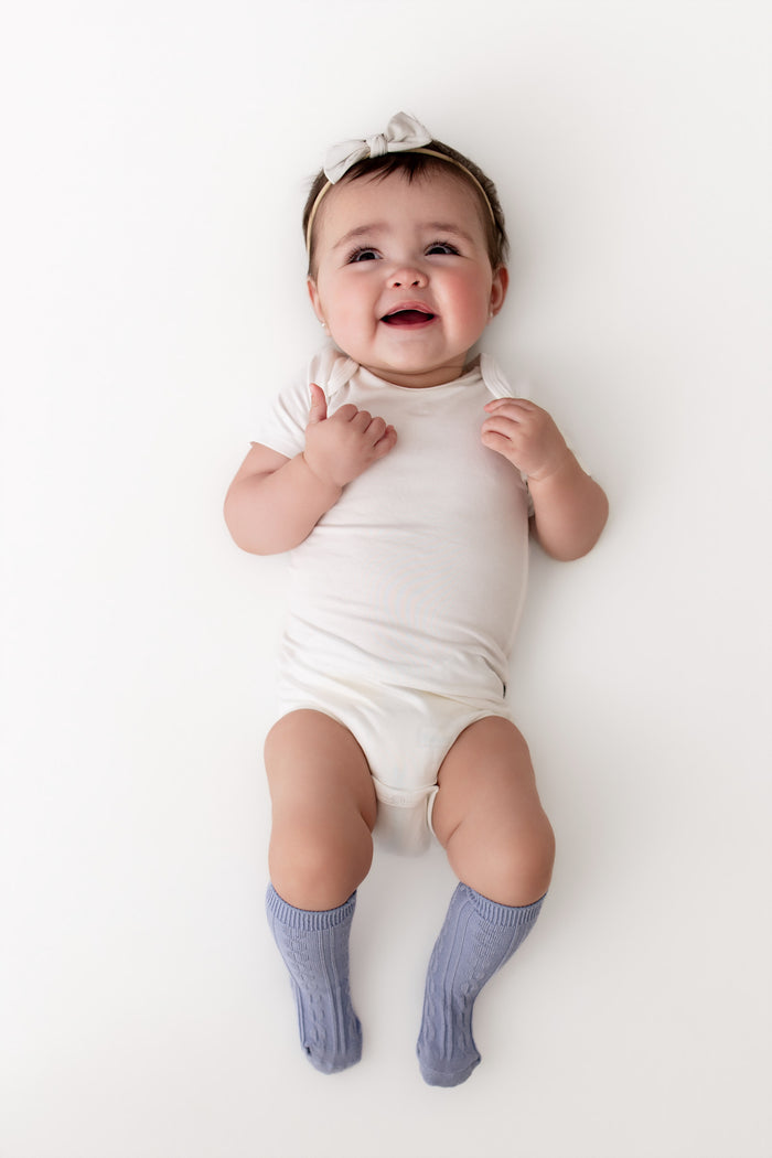A smiling baby lying on a white background, wearing a white short-sleeve bodysuit, a white bow headband, and light blue ribbed socks.