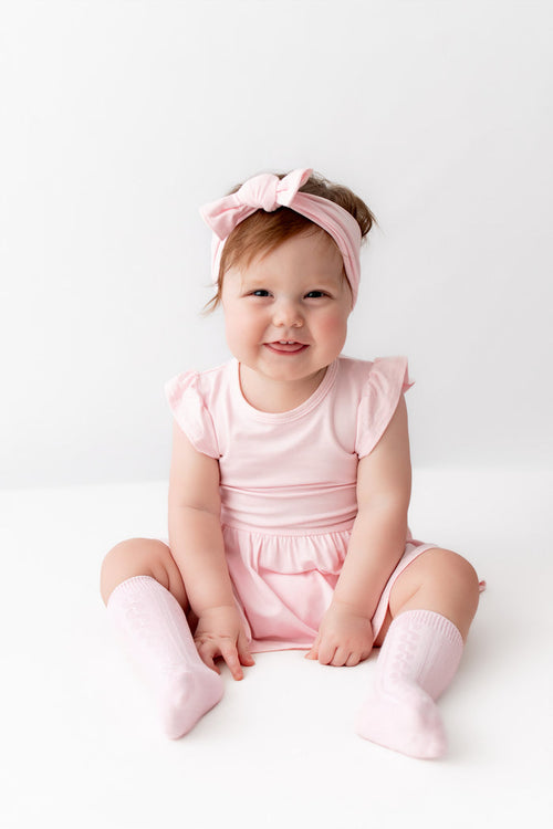 A smiling toddler girl sitting on a white studio floor, wearing a light pink ruffle-sleeve dress, matching pink knee-high socks, and a pink bow headband.