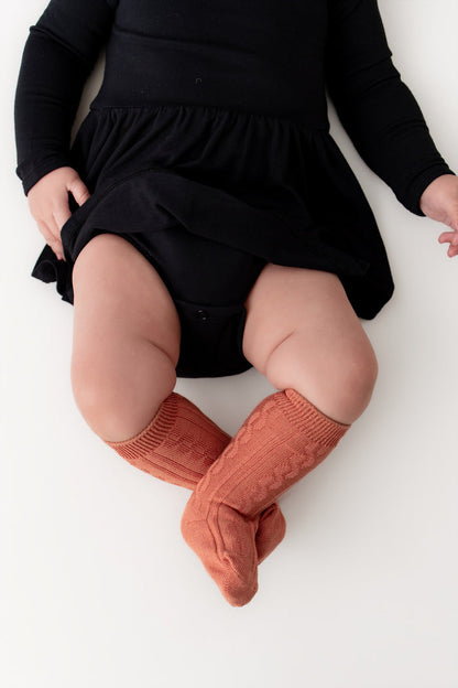 A baby wearing a black long-sleeve dress and rust-colored ribbed knit socks, lying on a white background.