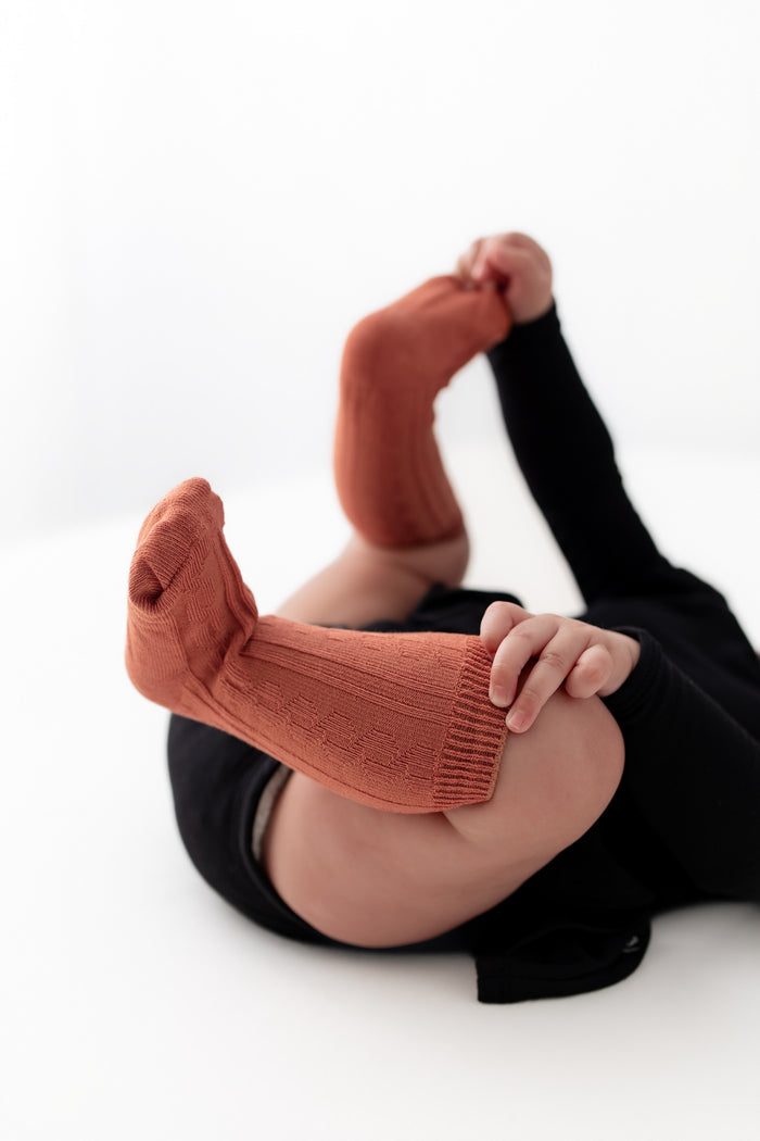 A person lying on a white floor wearing ribbed knit socks in a muted terracotta or rust color, paired with black pants. The person is holding their feet and legs bent upward.