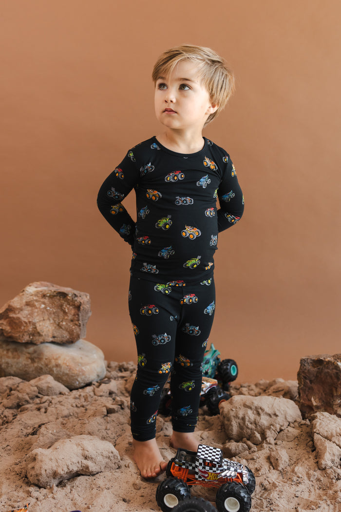 Boy wearing black Long Sleeve Pajamas in Monster Truck print standing on sand and rocks with toy trucks