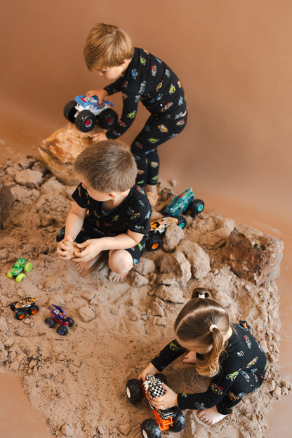 Kids playing with toy trucks wearing black Long Sleeve Pajamas, zippered romper and short sleeve toddler pajama set in Monster Truck print on sand and rocks with toy trucks around