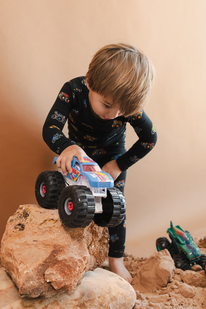Boy playing with toy truck wearing Black Long Sleeve Pajamas in Monster Truck print on sand and rocks with toy trucks