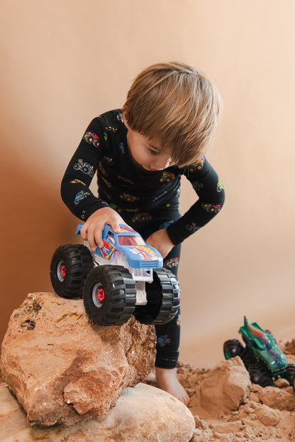 Boy playing with toy truck wearing Black Long Sleeve Pajamas in Monster Truck print on sand and rocks with toy trucks