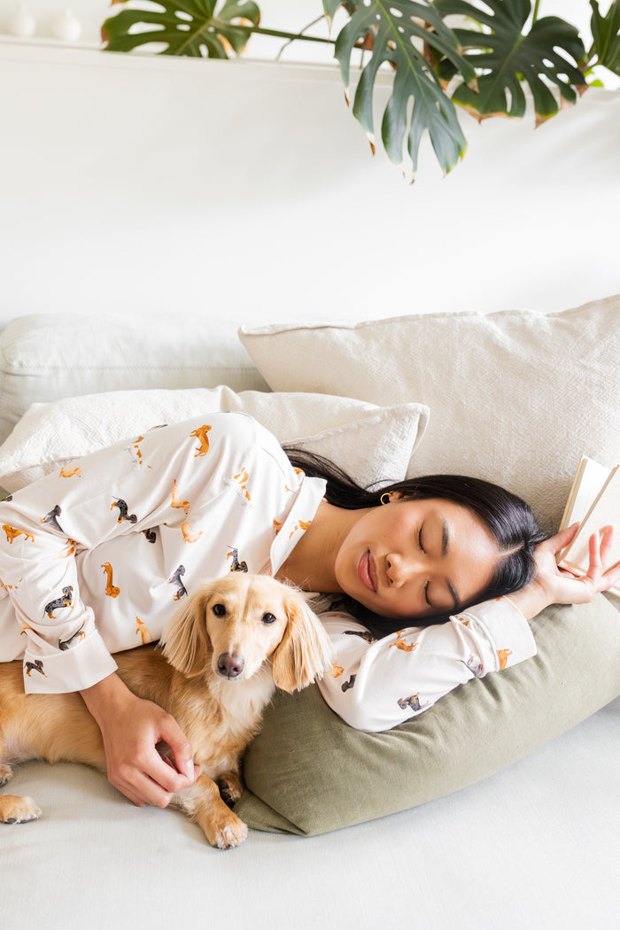 Woman in dachshund dog patterned pajamas lying on a couch with a small dog, surrounded by plants