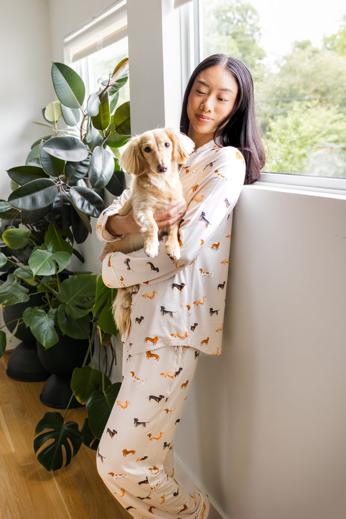 Model wearing Long-Sleeved Women's Pajama Set in Dachshund print standing against a wall near a tree holding a Dachshund dog