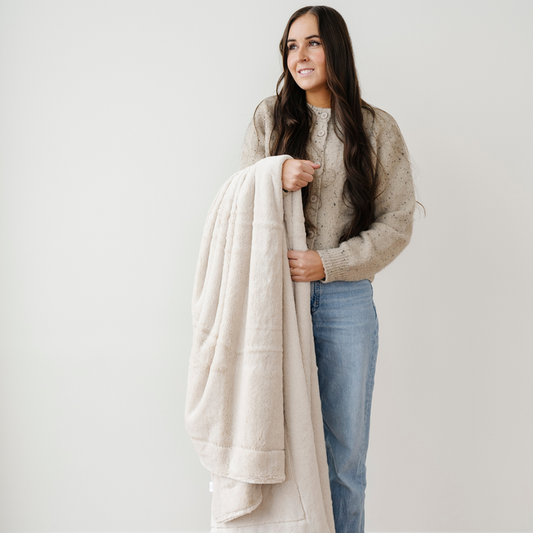 A woman smiles and holds a cream blanket that is a lush blanket. This soft blanket is a luxury blanket and a Saranoni blanket. Perfect as a home decor staple, for gifting, or for relaxation, this extra large throw blanket is sure to elevate and bring everyday luxury to your home. Enjoy this cozy blanket for years to come.