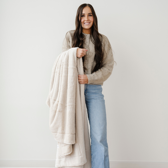 A woman smiles and holds an extra large blanket that is a buttermilk color and is also a lush blanket. This soft blanket is a luxury blanket and a Saranoni blanket. Perfect for gifting, relaxing, or as a home decor staple. Enjoy this cozy blanket for years to come.