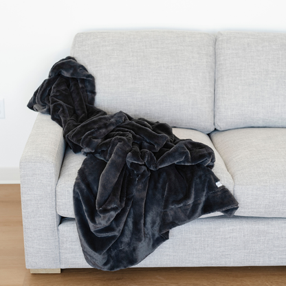 A charcoal blanket that is a lush blanket is draped over a gray couch. This soft blanket is a luxury blanket and a Saranoni blanket. This cozy blanket has a luxurious texture and is great for relaxing, gifting, or as a home decor item. Enjoy this extra large blanket for years to come.