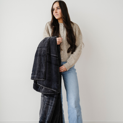 A woman holds a charcoal lush blanket that is a Saranoni blanket. This soft blanket is also a luxury blanket and an extra large blanket. Gift this cozy blanket to someone, use it for relaxing, or add it to your home as a home decor staple. Enjoy this luxury blanket for years to come.