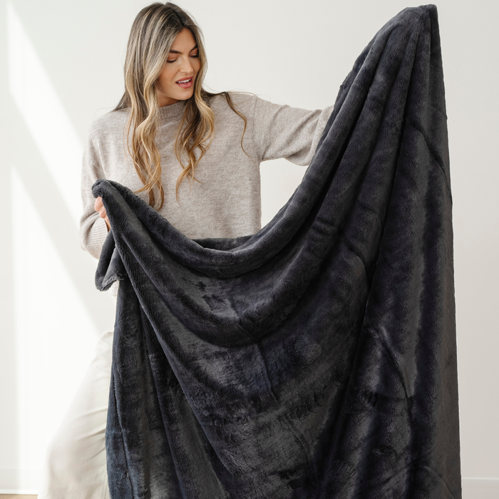 A woman holds up a charcoal blanket that is an extra large blanket. This soft blanket is a lush blanket and also a Saranoni blanket. Use this luxury blanket for gifting, relaxing, or as an addition to your home decor. Enjoy this cozy blanket for years to come.