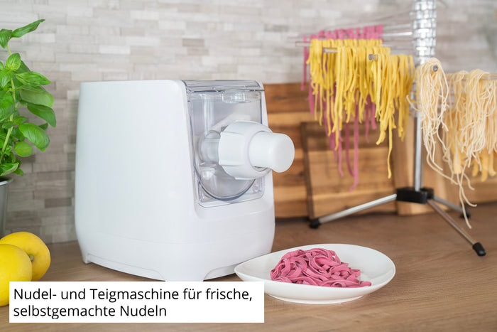 Die Unold Nudelmaschine Nora, eine weiße Nudel- und Teigmaschine, steht auf einer Küchentheke neben violetten Nudeln, gelben Nudeln auf einem Trockengestell, Zitronen und einem Holzbrett. Ein deutscher Text beschreibt die Maschine.