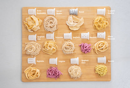 Die Unold Nudelmaschine Nora verfügt über 15 Nudelsorten, jede mit deutscher Beschriftung und den entsprechenden Aufsätzen auf einem Holzbrett angeordnet, und zeigt Formen wie Spaghetti, Fettuccine, Ravioli und Makkaroni.