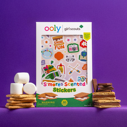 A set of OOLY x Girl Scouts S’more Scented Stickers surrounded by s'mores ingredients on a purple background
