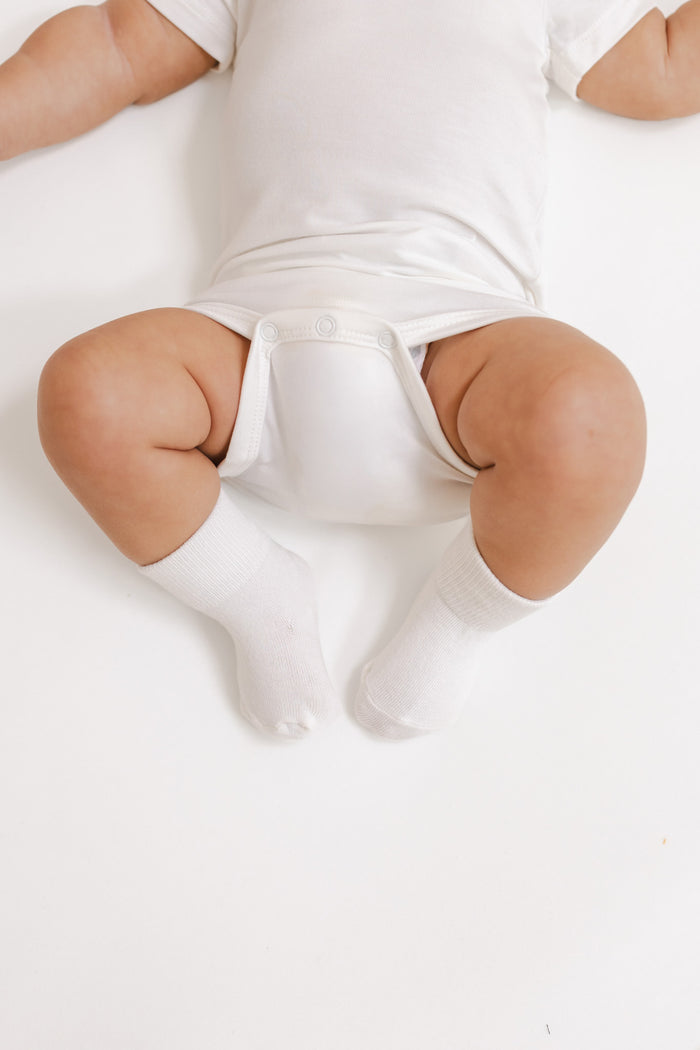 A baby lying on their back wearing a white short-sleeve bodysuit and white socks on a white background.