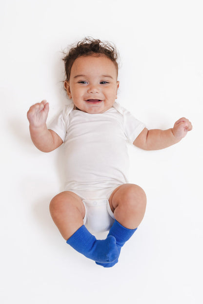 A smiling baby lying on their back wearing a white short-sleeve bodysuit and bright blue socks, photographed against a clean white background.