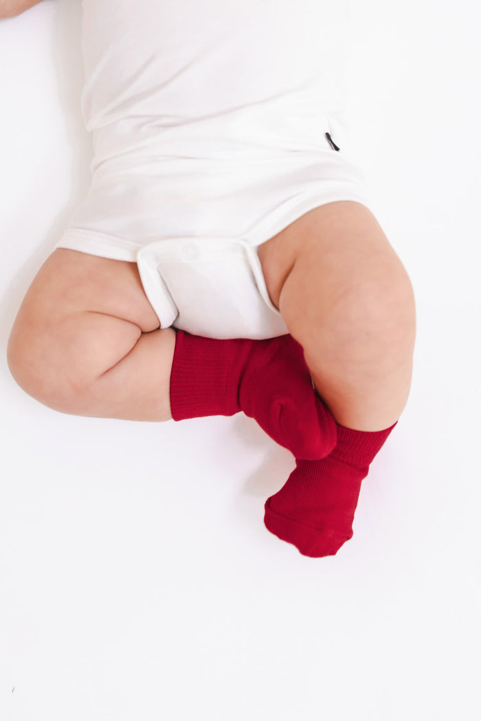 A close-up of a baby's legs wearing bright red knit socks and a white bodysuit, lying on a white surface.