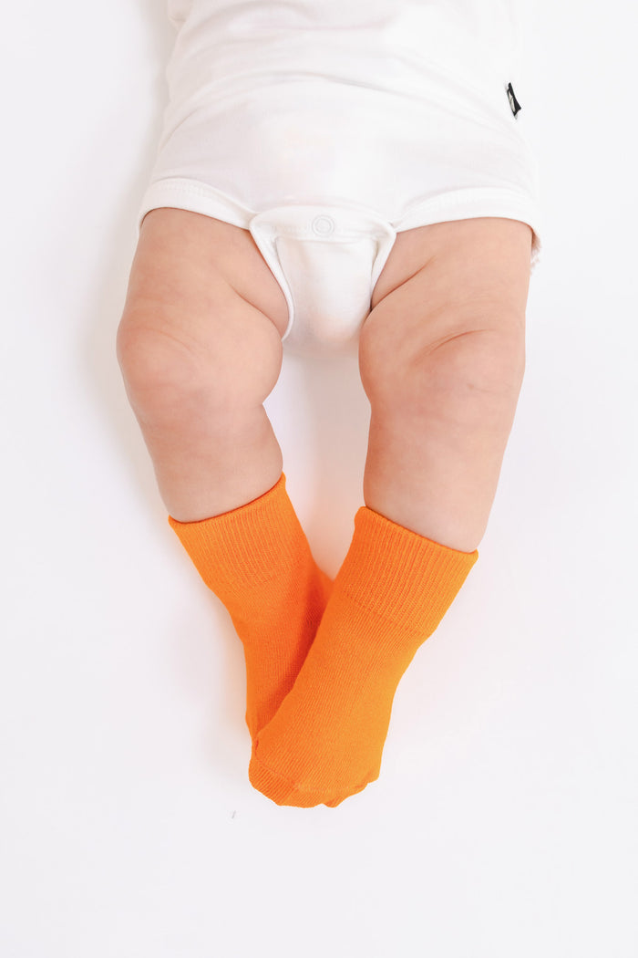 Bright orange baby socks worn by a baby in a white bodysuit, photographed from above on a clean white background.