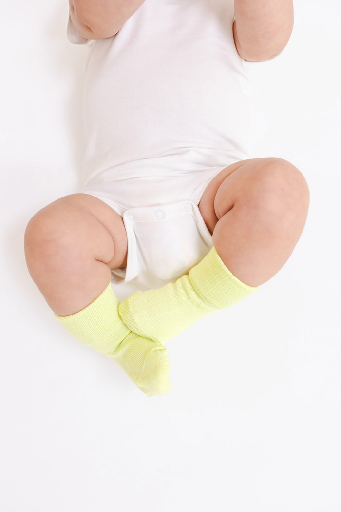 A baby wearing bright yellow-green knee-high socks and a white bodysuit, photographed from above on a plain white background.