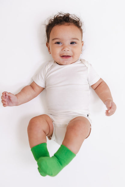 A baby lying on their back wearing a white short-sleeve bodysuit and bright green socks, photographed against a clean white background.