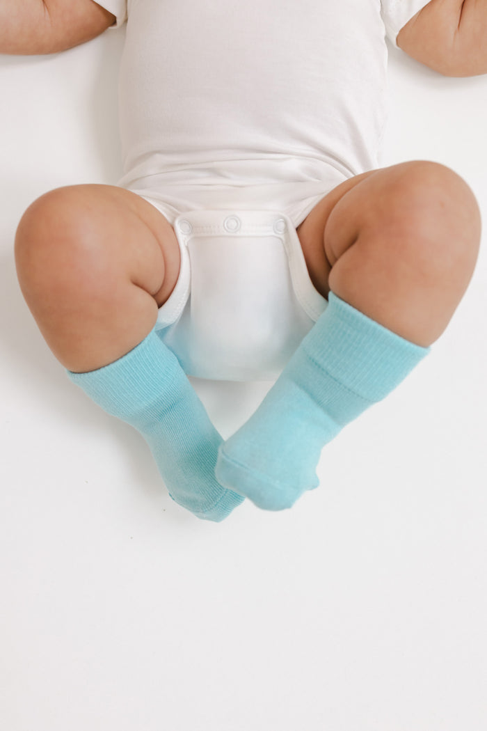 A baby wearing light blue ribbed socks with a white bodysuit and diaper, lying on a white surface.