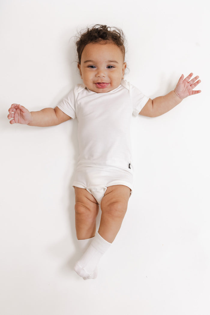A smiling baby lying on their back wearing a white short-sleeve bodysuit, white socks, and a white diaper, set against a seamless white background.