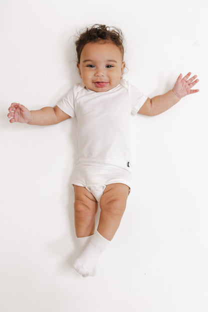 A smiling baby lying on their back wearing a white short-sleeve bodysuit, white socks, and a white diaper, set against a seamless white background.