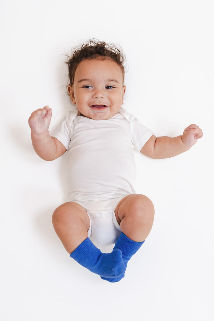 A smiling baby lying on their back wearing a white short-sleeve bodysuit and bright blue socks, photographed against a clean white background.