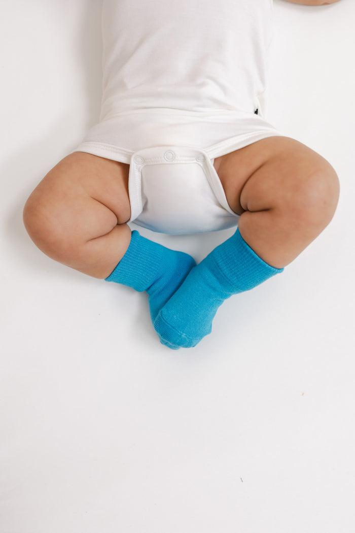 A baby lying on their back wearing bright turquoise blue socks and a white bodysuit, photographed from a top-down angle on a white background.
