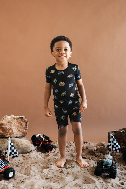 Boy wearing black Short Sleeve Pajamas in Monster Truck print standing on sand and stones with toy trucks