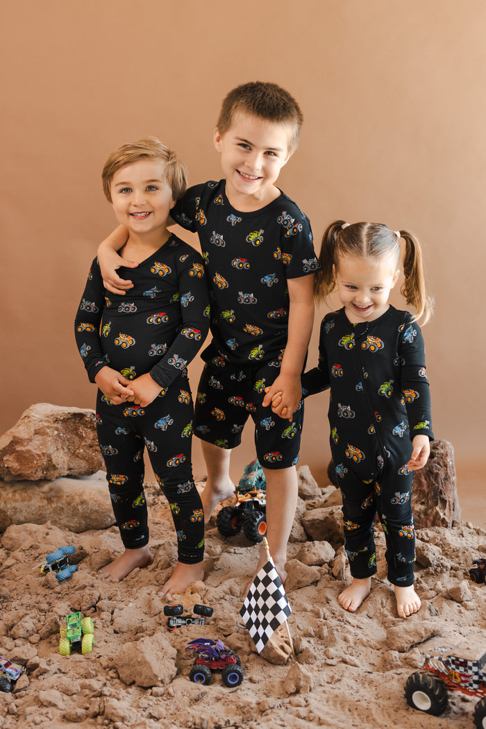 Kids wearing black Long Sleeve Pajamas, zippered romper and short sleeve toddler pajama set in Monster Truck print standing on sand and rocks with toy trucks around