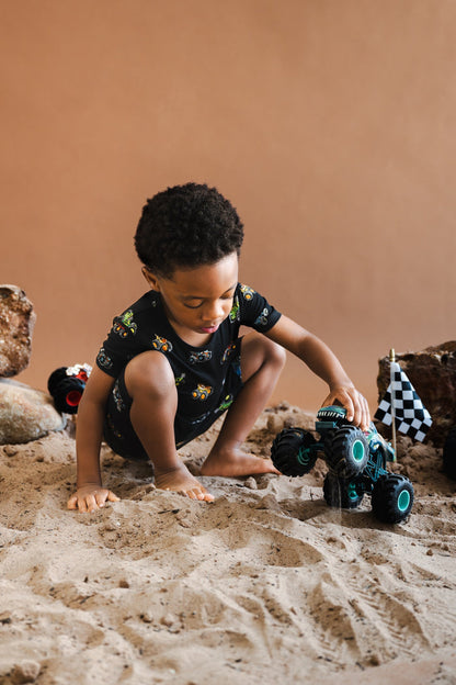 Boy playing with toy truck on sand and rocks wearing black Short Sleeve Pajamas in Monster Truck print