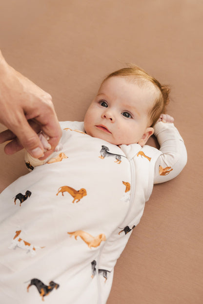 Baby holding a person's hand wearing a Beige Sleep Bag with Dachshund print on a brown background