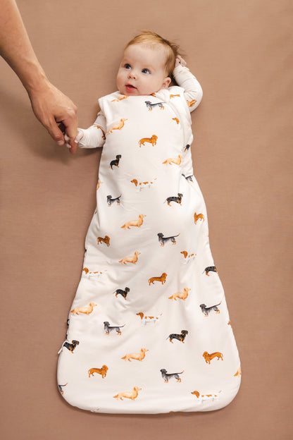 Baby in Beige Sleep Bag with Dachshund print holding a person's hand on a brown background