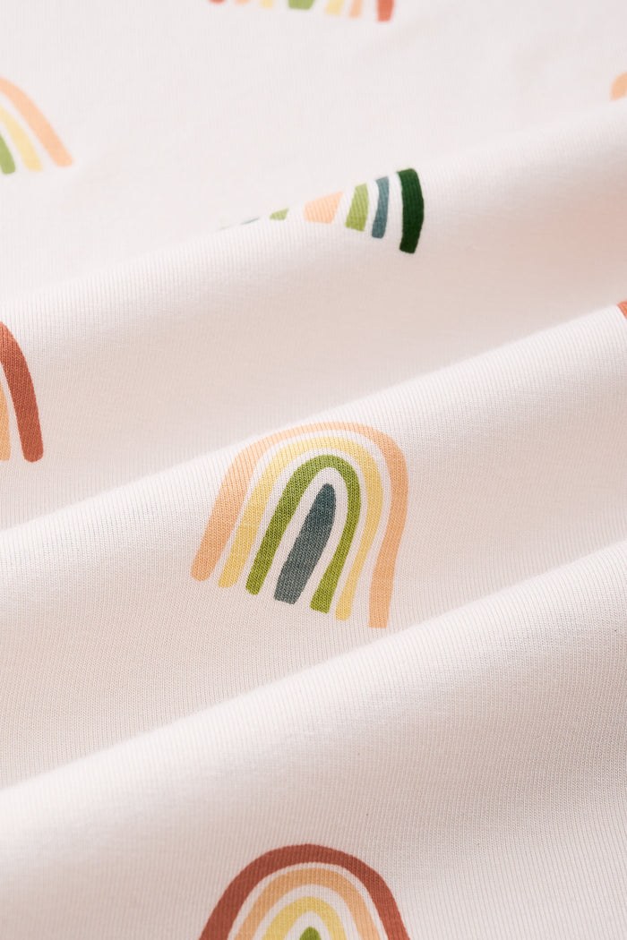 An all-over print featuring alternating rows of boho style rainbows in alternating directions over a light beige background color called Vanilla. 

One row of rainbows has an outermost arc colored in a dark cool green, followed by a cool sage green, then a light olive green, and finished with an inner core colored in a light apricot. 

Another row of of rainbows has an outermost arc colored in a burnt orange called Apricot, followed by a light peach, then a light yellow called Lilikoi, and finis