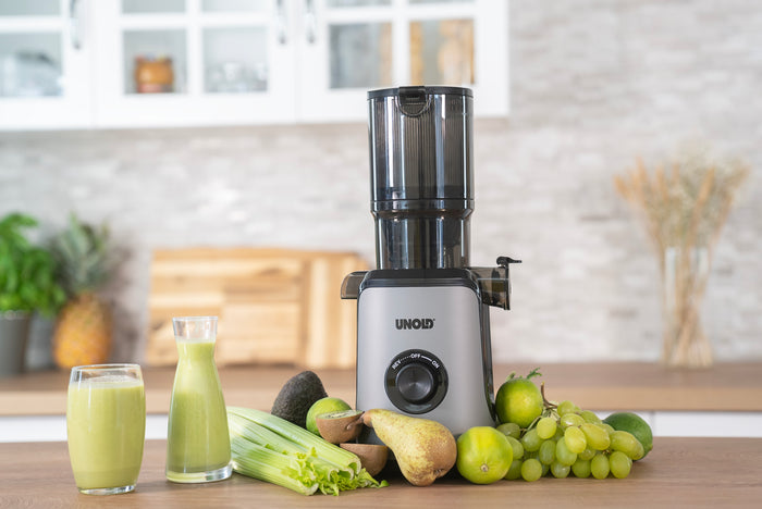 Der Unold Slow Juicer Sam steht auf einer Küchentheke, umgeben von Trauben, Sellerie, Birnen, Limetten, Avocado und Äpfeln, davor stehen zwei Gläser mit grünem Saft.