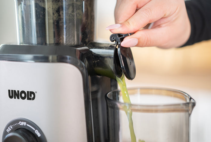 Eine Person öffnet den Ausguss des Unold Slow Juicer Sam und lässt frischen grünen Saft in einen darunter stehenden Glasbehälter fließen.
