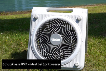 Ein weißer, quadratischer Unold Solarventilator Camping steht auf einer Wiese am Wasser. In der Mitte ist das Unold-Logo zu sehen. Text: Schutzklasse IPX4 – ideal bei Spritzwasser.