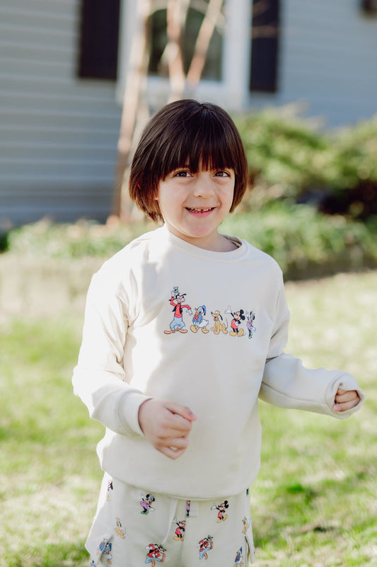 Child wearing a white sweatshirt with cartoon mickey character designs outdoors.