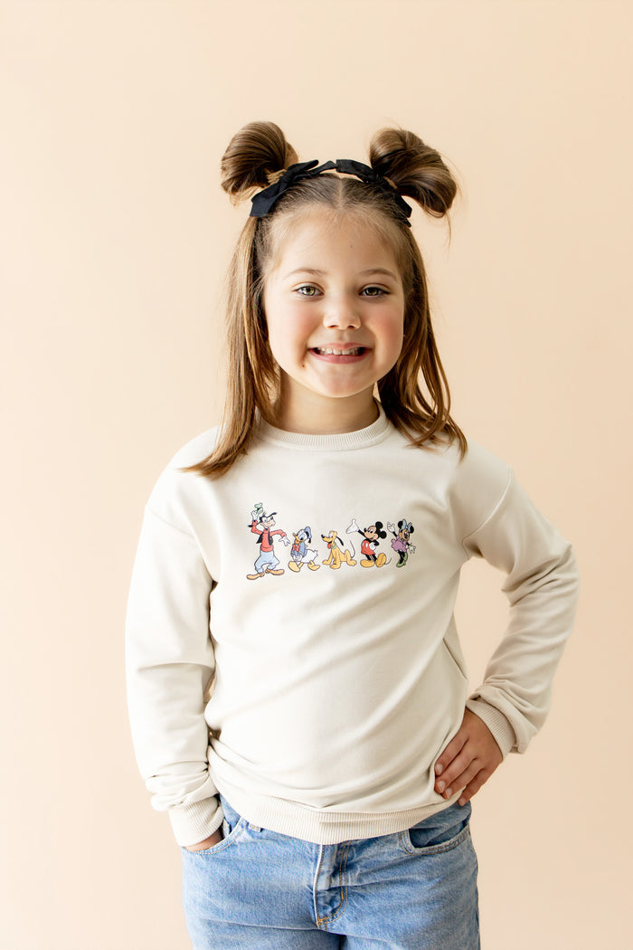 Young girl in cream long sleeve crewneck sweatshirt with mickey characters graphic across chest of sweatshirt