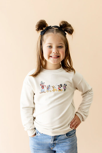 Young girl in cream long sleeve crewneck sweatshirt with mickey characters graphic across chest of sweatshirt