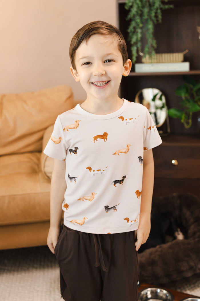 Boy standing in a living room wearing a Beige Toddler Basic Tee in Dachshund print over brown Drawstring