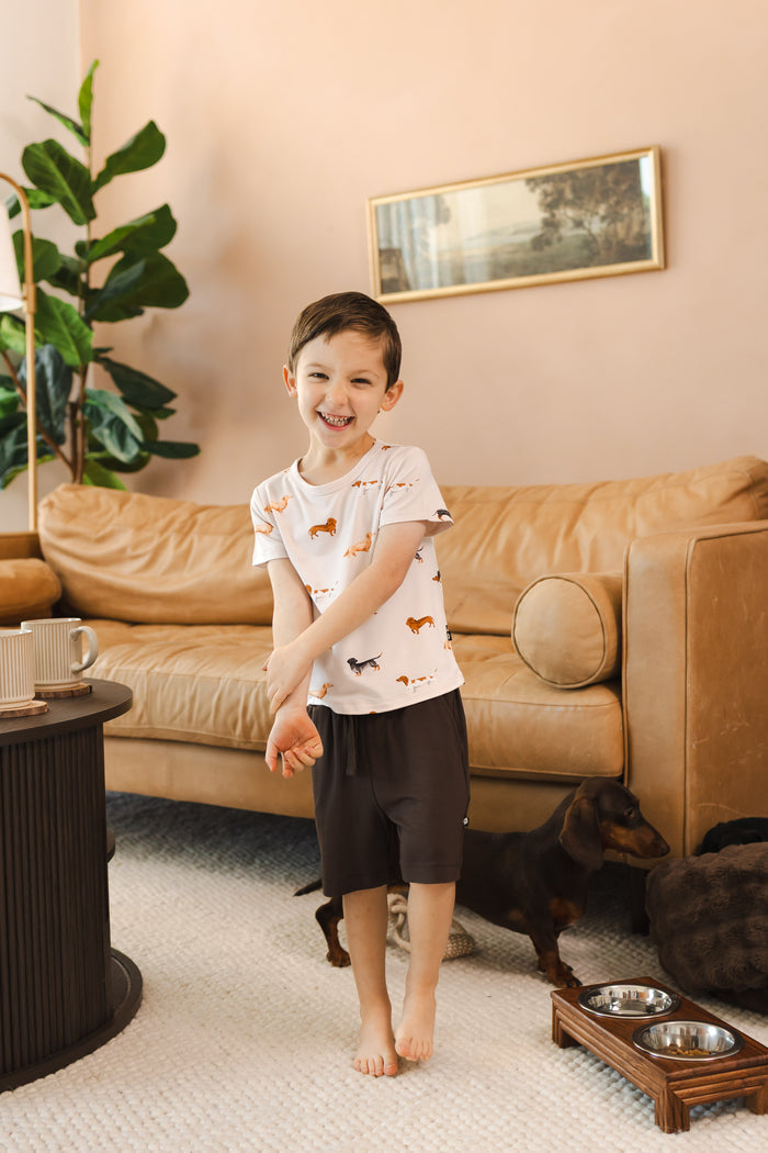 Boy standing in a living room smiling in a Beige Toddler Basic Tee in Dachshund print over brown Drawstring with a Dachshund dog.