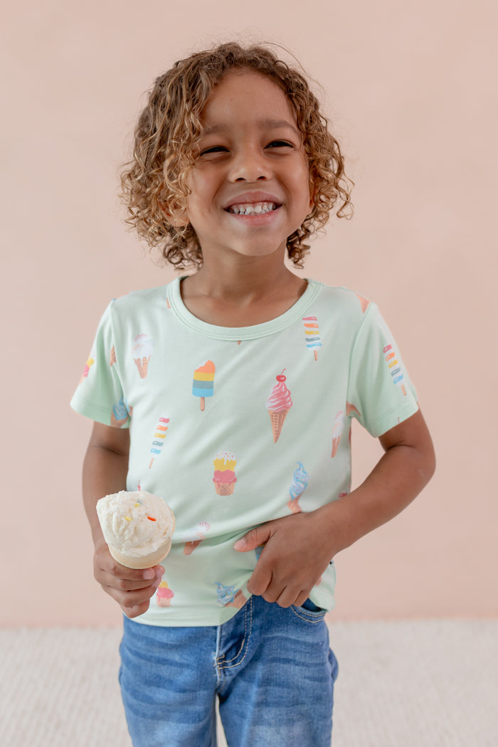Child smiling in Toddler Basic Tee in Mint Ice Cream print over denim pants holding an ice cream cone on a beige background