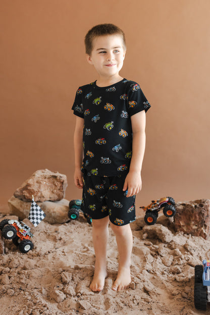Boy standing on sand and rocks with toy trucks wearing Black Toddler Basic Tee and Drawstring short in Monster Truck print