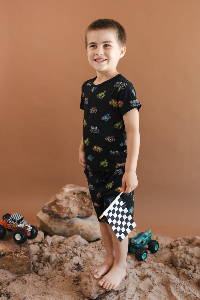 Boy smiling in Black Toddler Basic Tee and drawstring short in Monster Truck print standing on sand and rock with toy trucks