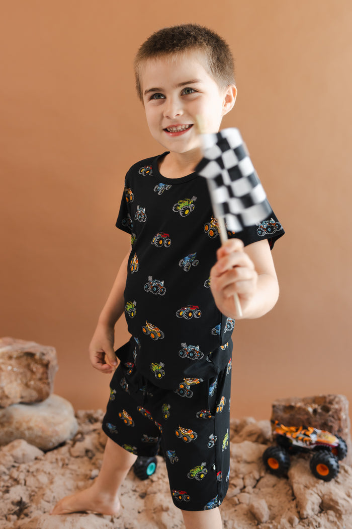 Boy holding a flag wearing Black Toddler Basic Tee and drawstring short in Monster Truck print standing on sand and rock with toy trucks